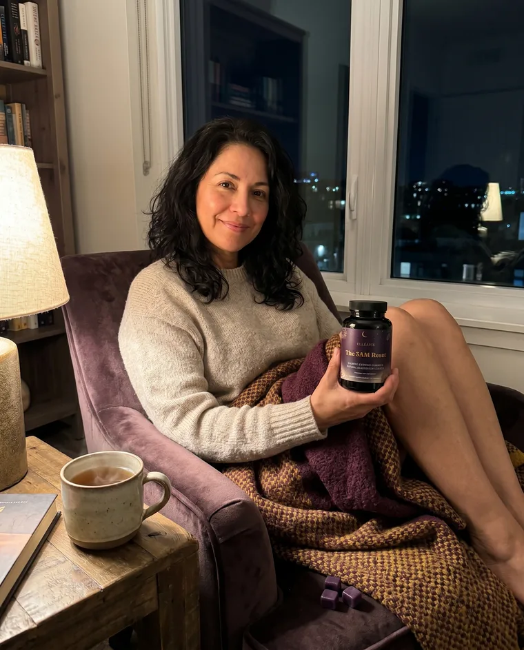 A woman relaxing in a reading chair at night with tea, holding The 3AM Reset.